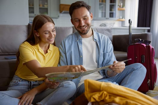 Happy loving couple reading paper map planning travel vacation during summer holidays. Cheerful man and woman sitting on floor front of stack of clothing prepared for trip