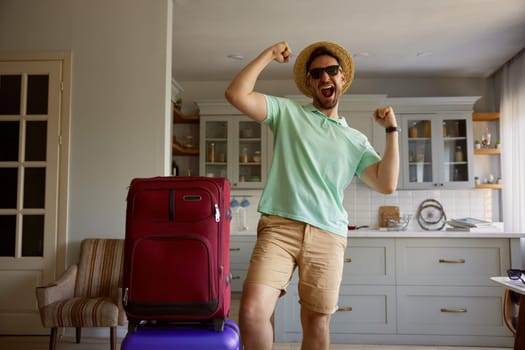 Happy man tourist wearing summer straw hat and sunglasses with luggage suitcases posing for camera over home interior background