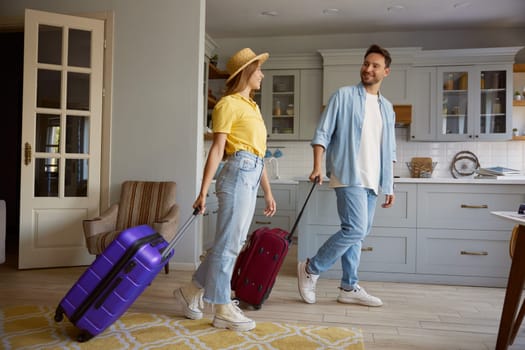 Happy smiling loving couple leaving home apartment with packed luggage ready for summer vacation trip, young man and woman feeling excitement and fun walking with suitcases