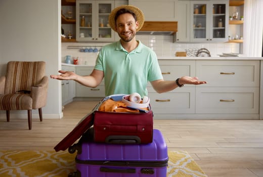 Happy man tourist wearing straw summer hat with packed stuff in travel suitcases posing for camera over indoor home interior. Vacation preparation and adventure time anticipation