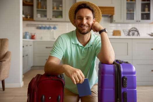 Portrait of happy man tourist wearing straw summer hat with packed stuff in travel suitcases posing for camera over indoor home interior. Vacation preparation and adventure time anticipation
