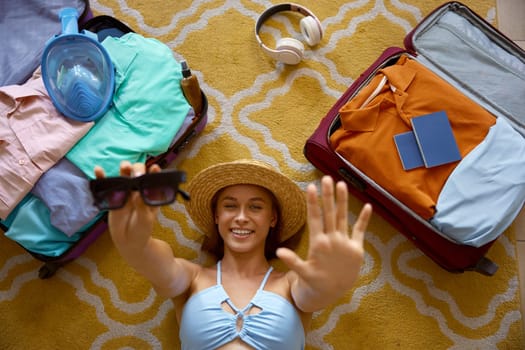 Happy young woman tourist wearing summer swimwear clothing posing for camera while lying on floor surrounded by packed travel suitcases with vacation stuff and passport