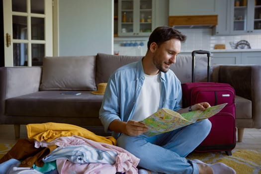 Young man tourist searching place of destination for travel reading paper map sitting on sofa in home living-room
