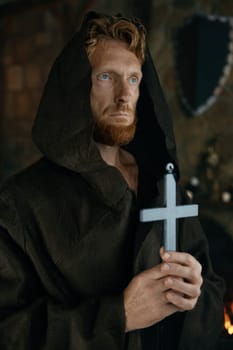 Medieval monk in robe praying holding silver cross in hand. Portrait of historic priest in monastery