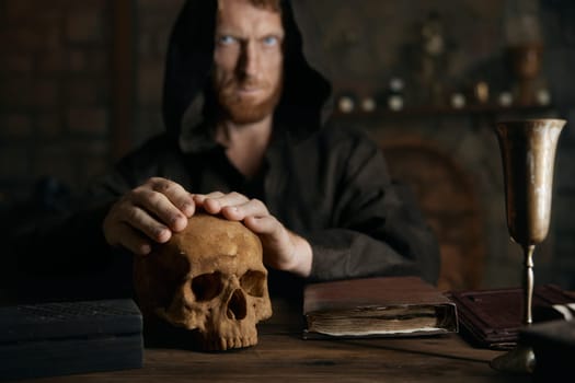 Portrait of mystical spooky medieval monk in black cassock putting hands on human skull prefroming spiritualistic seance or ritual. Fetal stage
