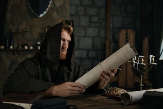 Religious man in hooded cape holding obsolete parchment scroll in hands sitting at wooden table of old mediaeval cathedral