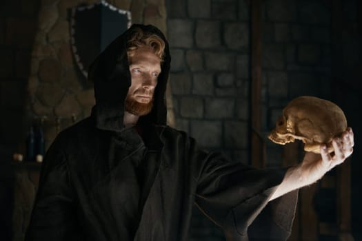 Serious medieval monk in hooded cloak looking at human skull in hands. Male priest sorcerer mysterious portrait