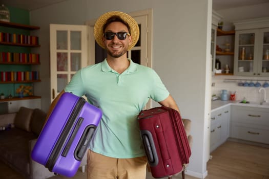 Portrait of happy man tourist wearing summer straw hat and sunglasses carrying packed luggage suitcases posing for camera over home interior background