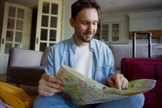Young man tourist searching place of destination for travel reading paper map sitting on sofa in home living-room