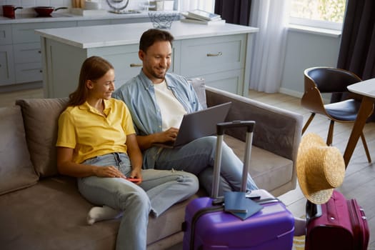 Happy loving couple planning honeymoon tour searching flight ticket or hotel for reservation on internet while sitting in home living-room with packed travel suitcases