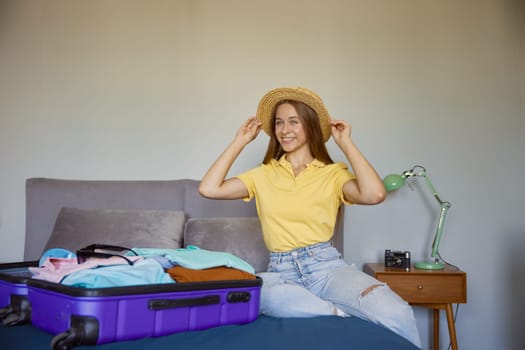 Joyful happy young woman rejoicing vacation time preparing suitcase luggage with clothes while sitting on bed at home