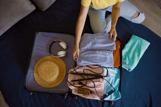 Unrecognizable woman traveler packing luggage preparing for summer vacation. Closeup view from above on female hands putting clothes and personal belongings into suitcase