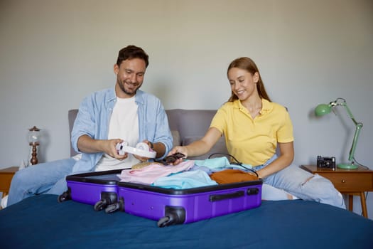Joyful married couple planning vacation preparing clothes for travel. Happy smiling guy and girl sitting front of opened suitcase on home bed