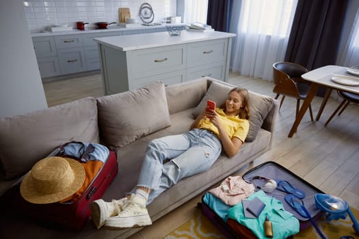 Young woman booking trip with phone while sitting on home sofa. Packing travel suitcases and planning travel vacation
