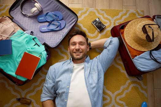 Young man dreaming about future vacation trip while lying on floor surrounded with opened suitcases filled personal belongings and clothes. Headshot with view from above