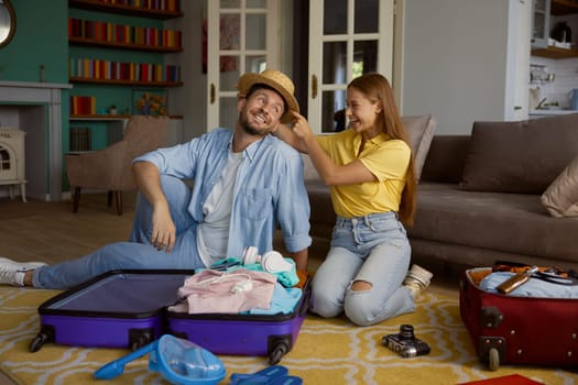 Overjoyed loving couple folding garment into travel suitcases while sitting on floor of home living-room feeling happy and playful