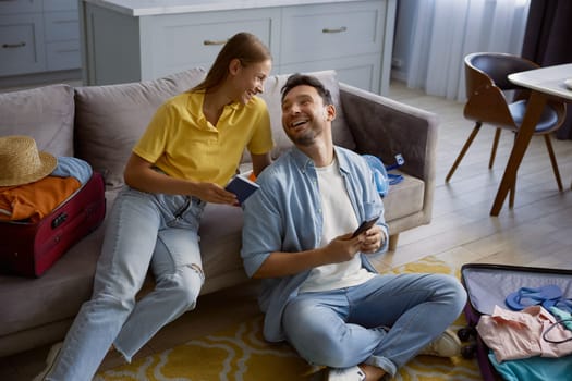 Joyful couple booking tickets or hotel room number at resort for vacation via mobile phone while preparing for travel at home