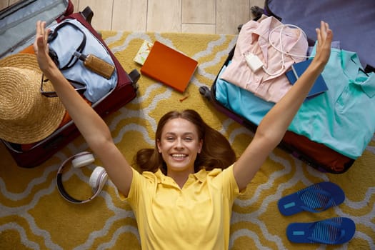 Satisfied excited woman with raised hands and opened travel bags around lying on floor rejoicing start time for summer vacation