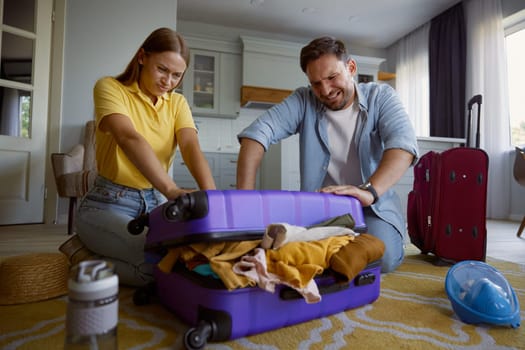 Young loving couple with frowning face trying to close overloaded travel bag during vacation preparation on summer weekends