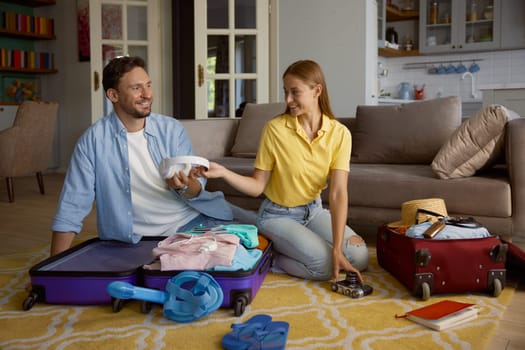 Loving couple folding garment into travel suitcases while sitting on floor of home living-room feeling happy and playful