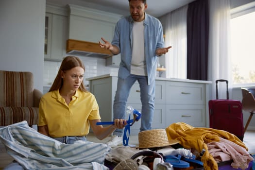 Couple quarreling because of clothing excess prepared for vacation. Angry man yelling on upset woman sitting on floor tying to put diving mask into travel luggage suitcase
