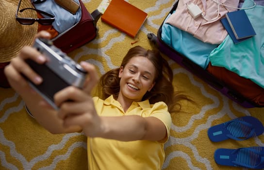 Young happy woman taking memory picture packing luggage for trip. Overhead view on pretty girl making selfie photo before going for travel lying on floor