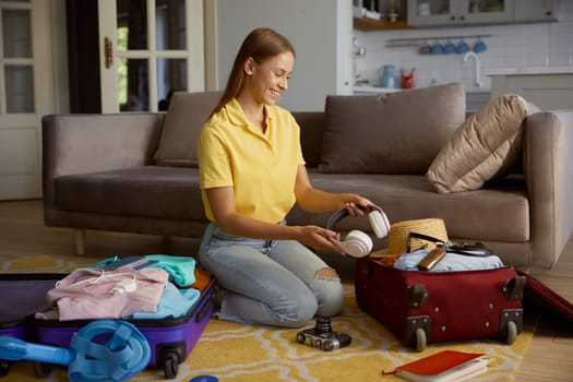 Young smiling woman tourist packing travel luggage putting headphones into bag with personal belongings