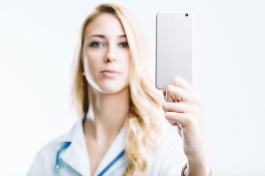 Young female doctor holding a smartphone with a blank screen, looking directly at the camera against a clean white background, exuding professionalism in her medical uniform
