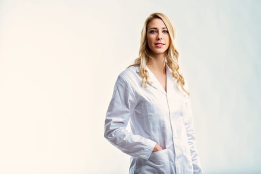 Young female doctor with long blond hair, wearing a white coat, stands confidently with her hands in her pockets against a white backdrop, exuding professionalism and expertise
