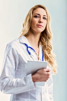 Young female doctor wearing a white coat and stethoscope is holding a digital tablet, looking directly at the camera with a confident expression