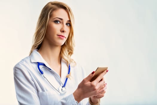 Young female doctor wearing lab coat and stethoscope, looking at camera while using smartphone with a confident expression, on a white background