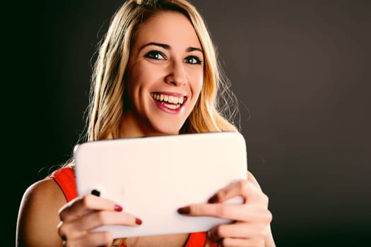 Young blonde woman with red dress holding a tablet and playing with it, smiling and looking at the camera, having fun with technology and entertainment