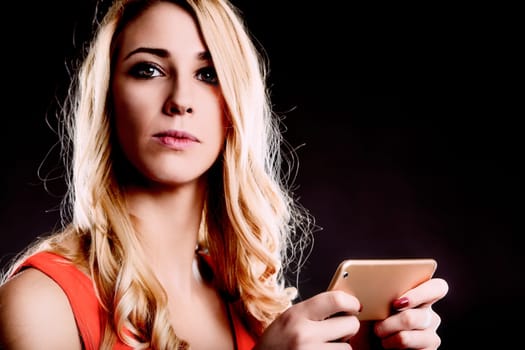 Studio shot of a serious young woman with long, wavy blond hair, wearing a sleeveless red top, holding a gold tablet computer in her hands, looking directly at the camera against a dark background