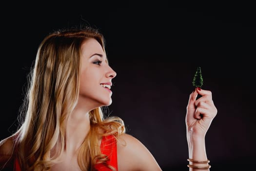 Studio shot of a happy young woman looking and smiling at a miniature christmas tree that she is holding between her fingers, against a black background