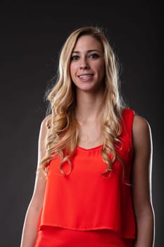 A blonde woman in an orange dress, caught in a half-length portrait. Gentle smile, relaxed arms, black backdrop, accented by light from the right