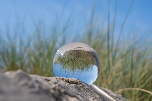 A glass ball lies in the sand of a dune by the sea, the dune grass and the blue sky are reflected in the ball