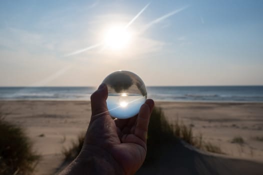 A hand of an  person holds a glass ball, the sea and the setting sun are reflected in the ball