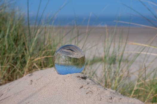 A glass ball lies in the sand of a dune by the sea, the dune grass and the blue sky are reflected in the ball