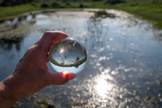A human hand is holding a glass ball, in the ball a small lake in green nature is reflected, the light of the sun is reflected in the water