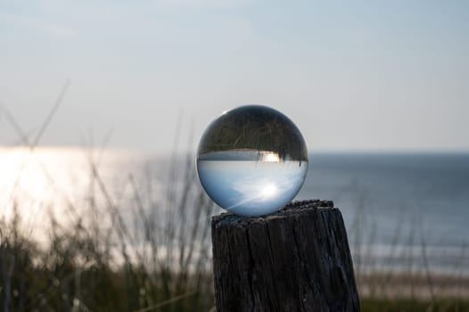 Glass ball lies on a wooden post, beach grass in the background, the sea, the sun and the blue sky are reflected in the ball