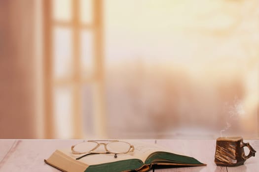 Books backgrounds. Closeup of an antique book with reading glasses on it and a brown clay mug on a light desk over a blurred windows. Education concept. Teachers day.