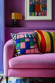 A vibrant pink sofa adorned with patterned cushions sits against a purple wall. A striking lamp and artwork add charm to the cozy space.
