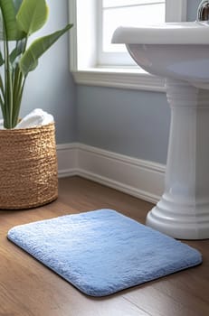 A soft blue bath mat rests on wooden flooring near a white sink and potted plant in a well-lit bathroom setting.