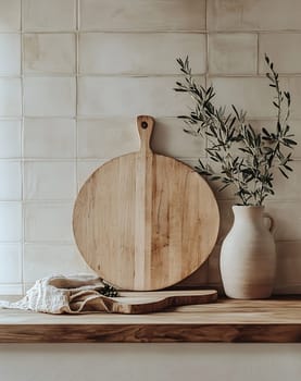 A wooden cutting board rests on a kitchen countertop beside a cream vase with green olive branches, creating a serene atmosphere.