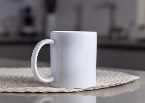 Closeup of 11 oz white blank coffee mug on grey kitchen table over blured modern kitchen grey interior, mockup. Template for your design or advertising with copy space