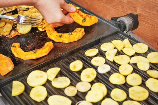 Bright orange butternut squash pieces grilled on electric grill, woman hands moving vegetables, blurred potato chips also cooked in foreground