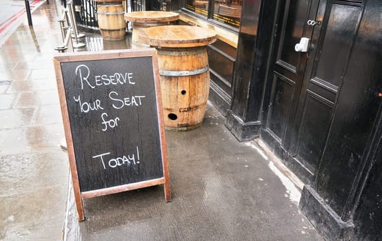 Black Chalkboard with "Reserve your seat for Today"" text in front of restaurant or pub black door. Wooden barrel tables standing on wet concrete at background"