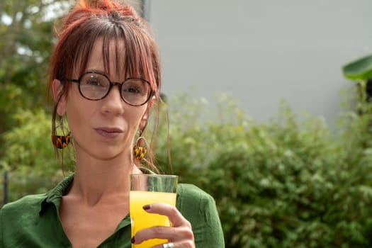 A woman with glasses and red hair is holding a glass of orange juice. She has a serious expression on her face