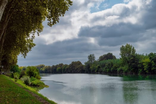 A river with trees on either side. The sky is cloudy and the water is calm. The scene is peaceful and serene. Peyrehorade, France