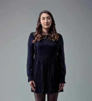 Young woman with long wavy brown hair, wearing a dark blue dress and black tights, standing against a neutral gray background, looking directly at the camera with a serious and confident expression